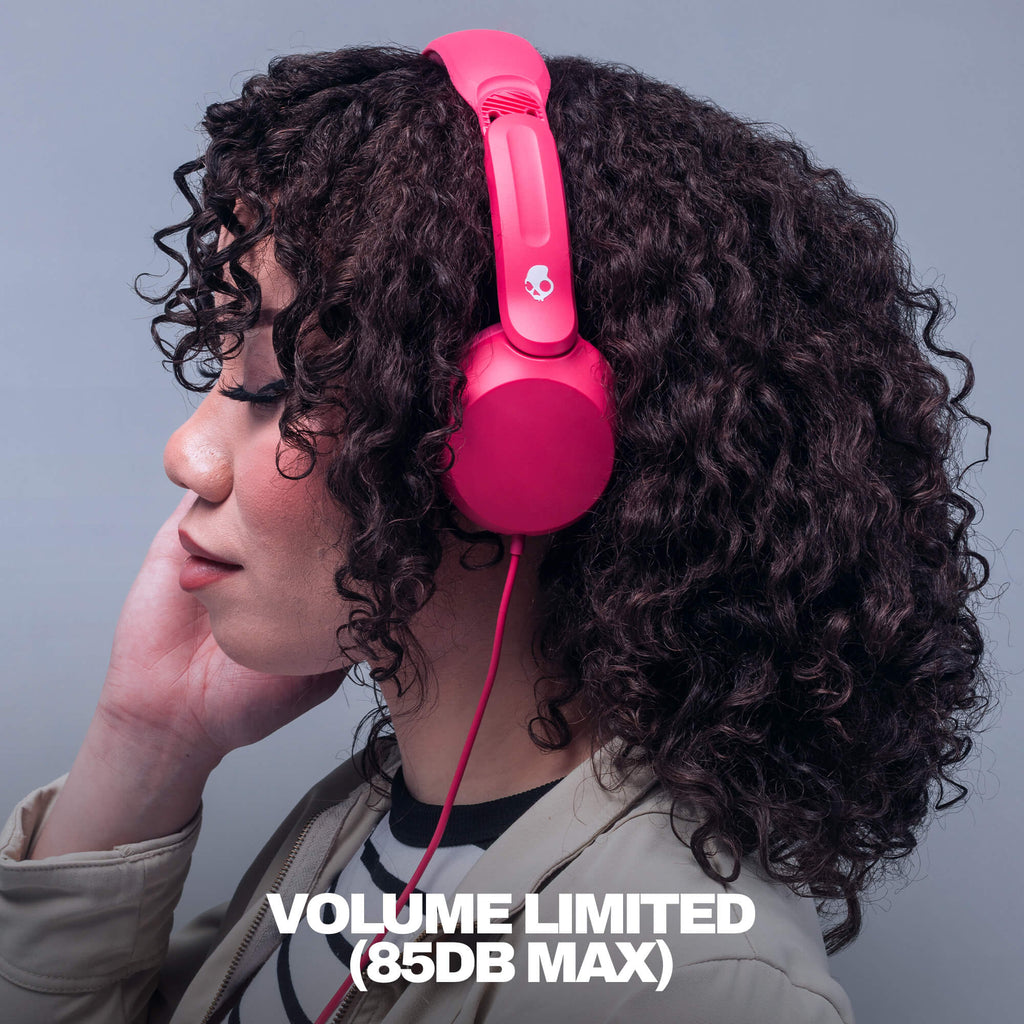 A person with curly dark hair wearing bright pink over-ear headphones, shown in profile against a plain gray background. White text at the bottom reads 'VOLUME LIMITED (85DB MAX)'. | Crazy Pink