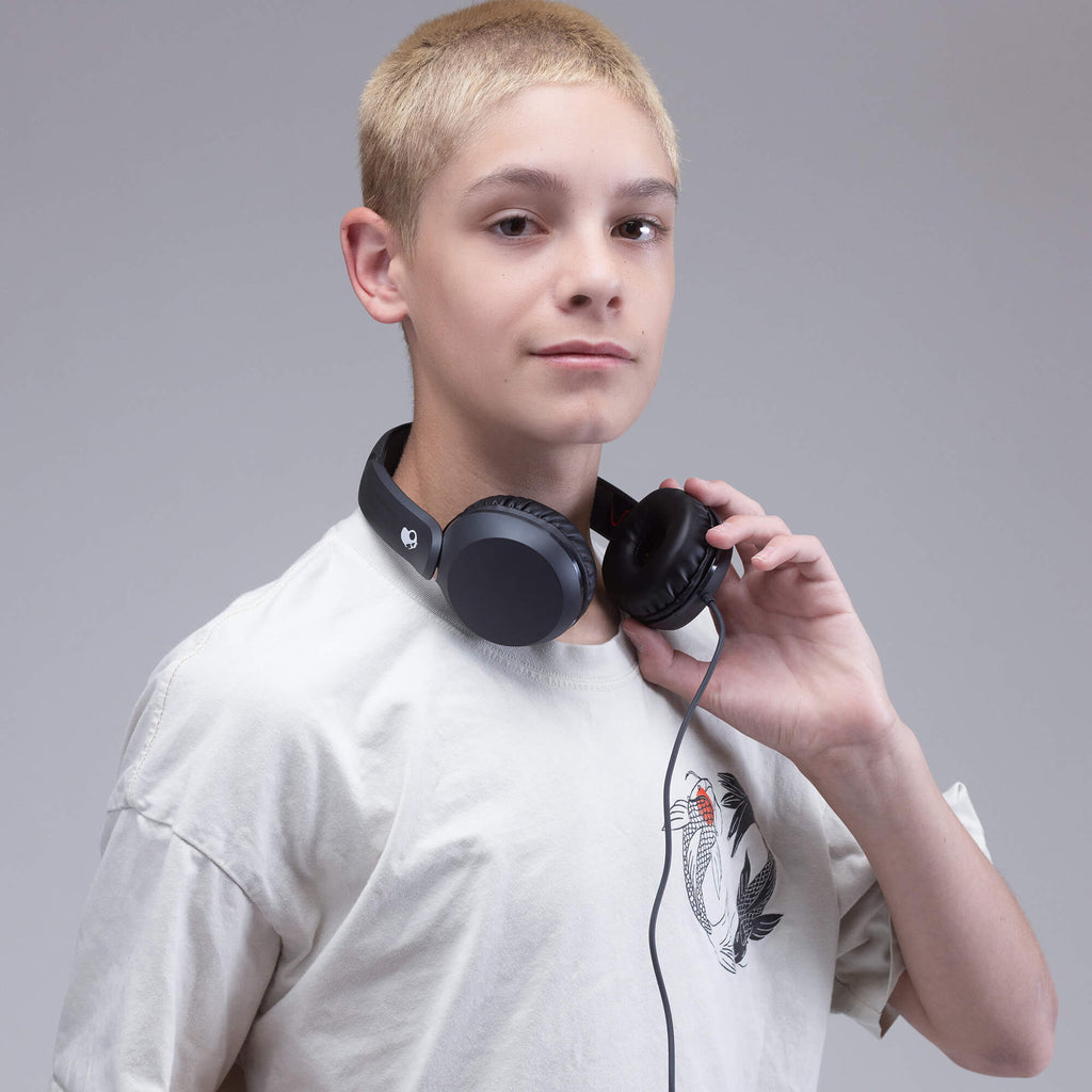 A young male model with short blond hair wearing a white t-shirt and holding a pair of black over-ear headphones around his neck, set against a solid gray studio background. | True Black
