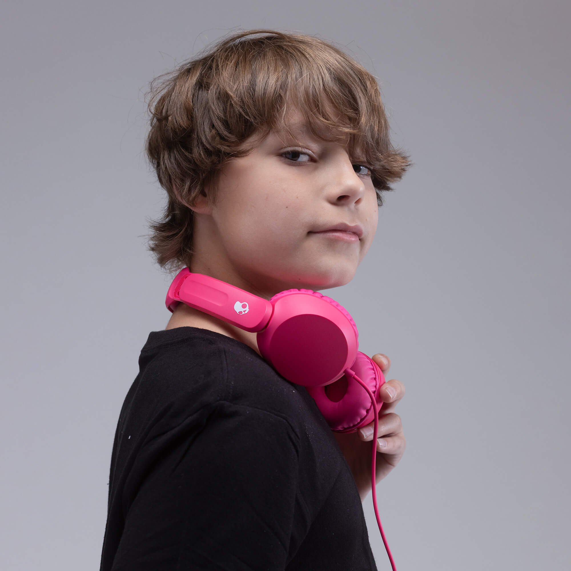 A child with brown hair wearing a black shirt, holding a pair of bright pink over-ear headphones around their neck against a solid gray background. | Crazy Pink