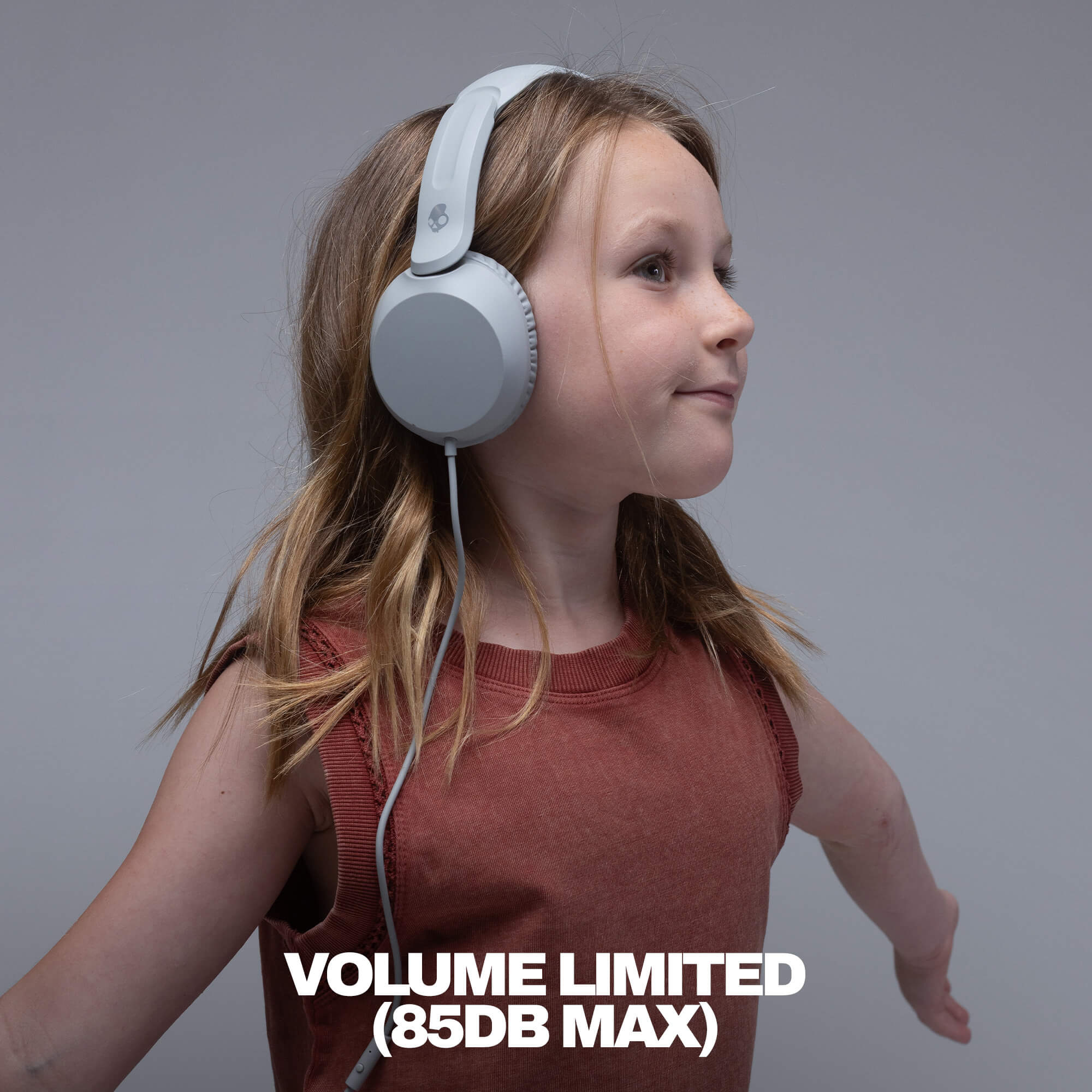 A young girl with blonde hair wearing light gray over-ear headphones and a rust-colored sleeveless top, shown in profile against a solid gray background with the text 'VOLUME LIMITED (85DB MAX)' at the bottom. | Cement