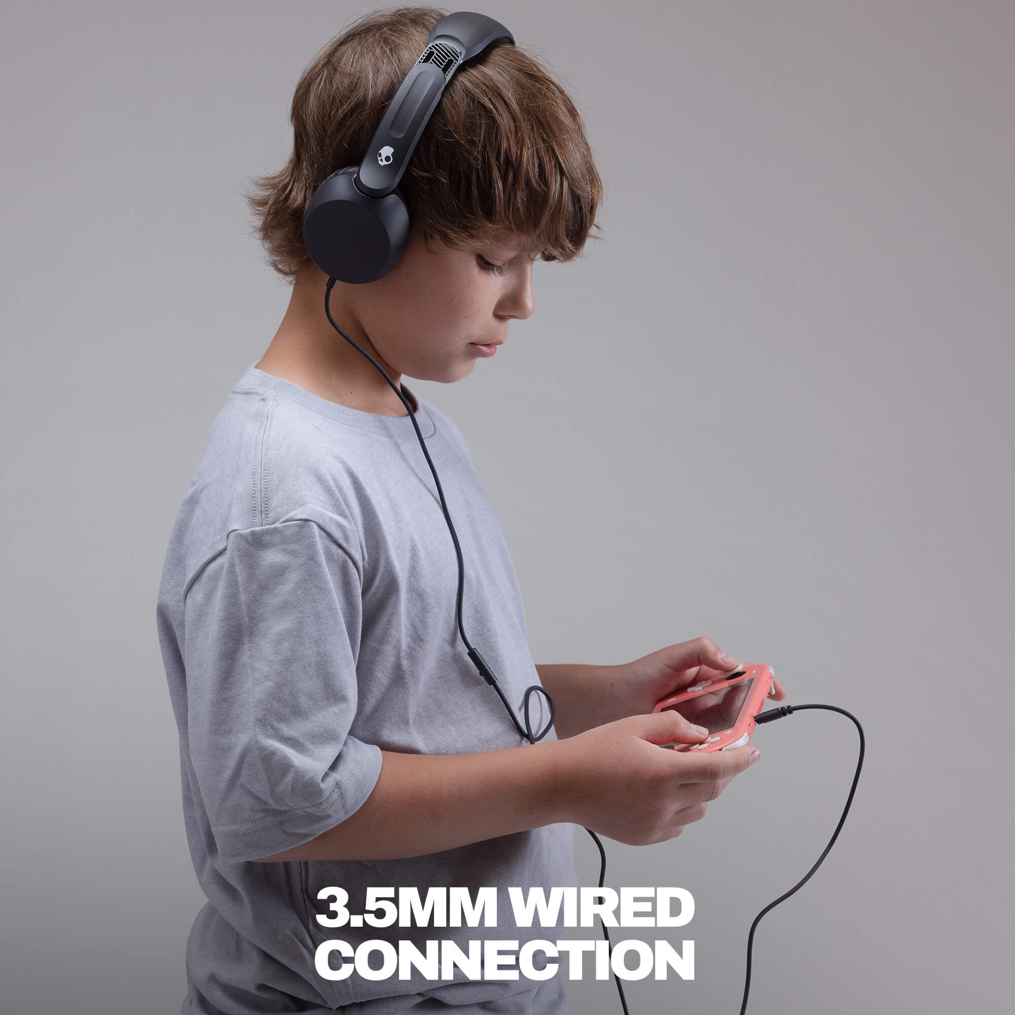 A child wearing black over-ear headphones and a light gray t-shirt, holding a pink smartphone with a black 3.5mm wired connection. | True Black