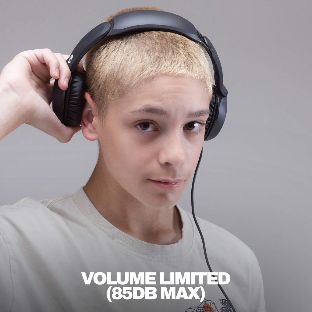 A person with short blond hair wearing black over-ear headphones and a light gray T-shirt, holding one earcup with one hand against a plain gray background. White text at the bottom reads 'VOLUME LIMITED (85DB MAX)'. Black over-ear wired headphones with cushioned ear pads and a padded headband, shown at an angle on a solid black background. A small white skull logo is visible on the headband. |True Black