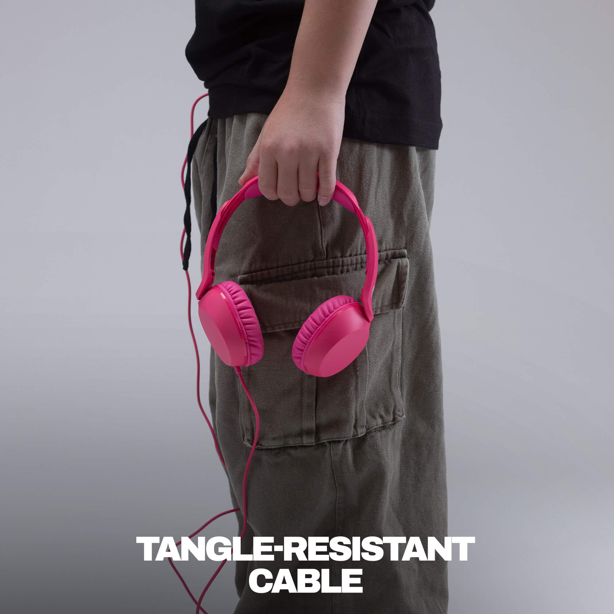 A person wearing a black t-shirt and olive cargo pants holds a pair of bright pink over-ear headphones with a pink cable. The text 'TANGLE-RESISTANT CABLE' is displayed at the bottom of the image against a solid gray background. | Crazy Pink