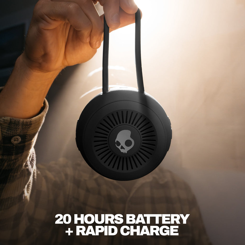Person holding a black portable speaker with a skull logo, text '20 Hours Battery + Rapid Charge' on a blurred background. | True Black