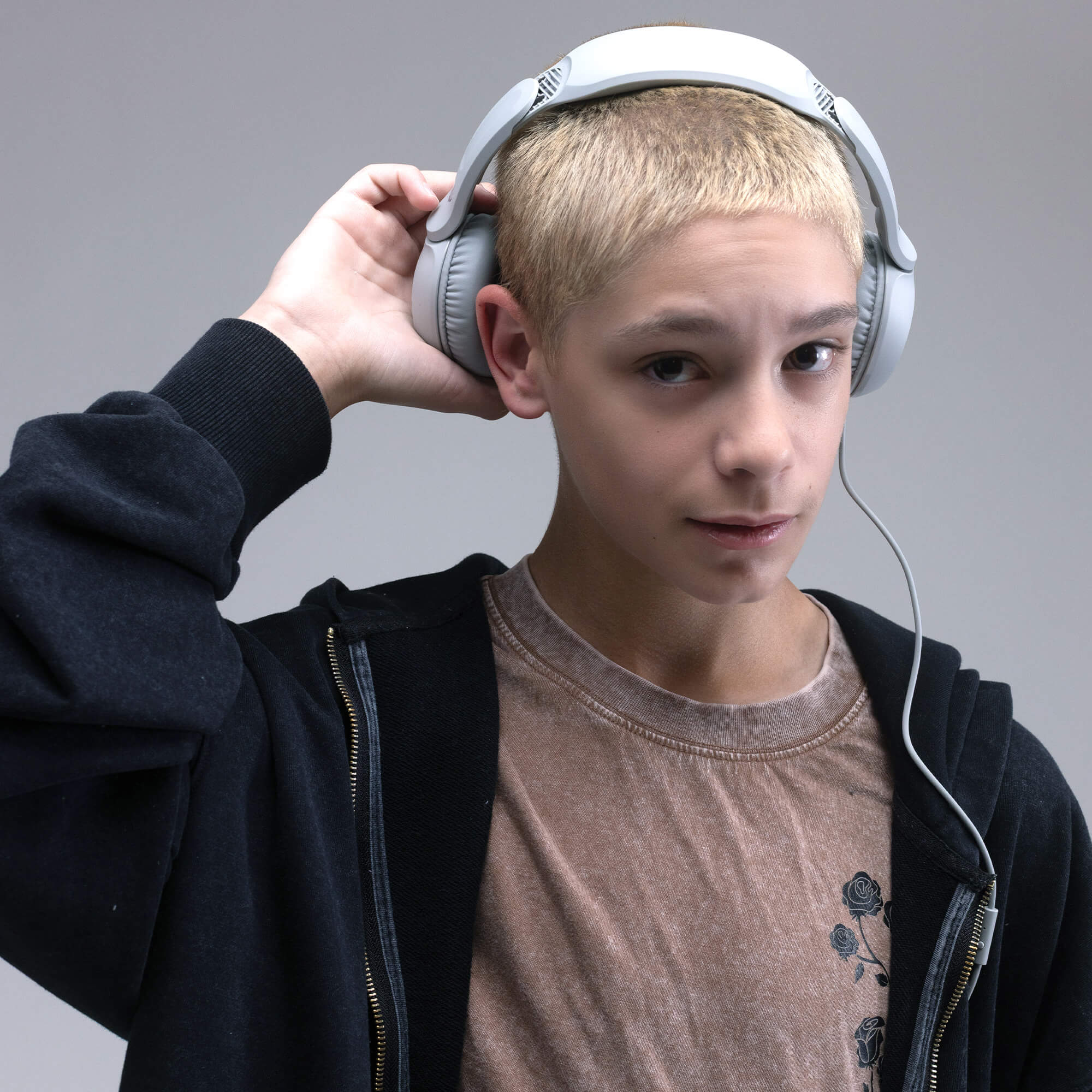 A young boy with short blond hair wearing light gray over-ear headphones, a black zip-up hoodie, and a brown t-shirt, set against a solid gray studio background. | Cement