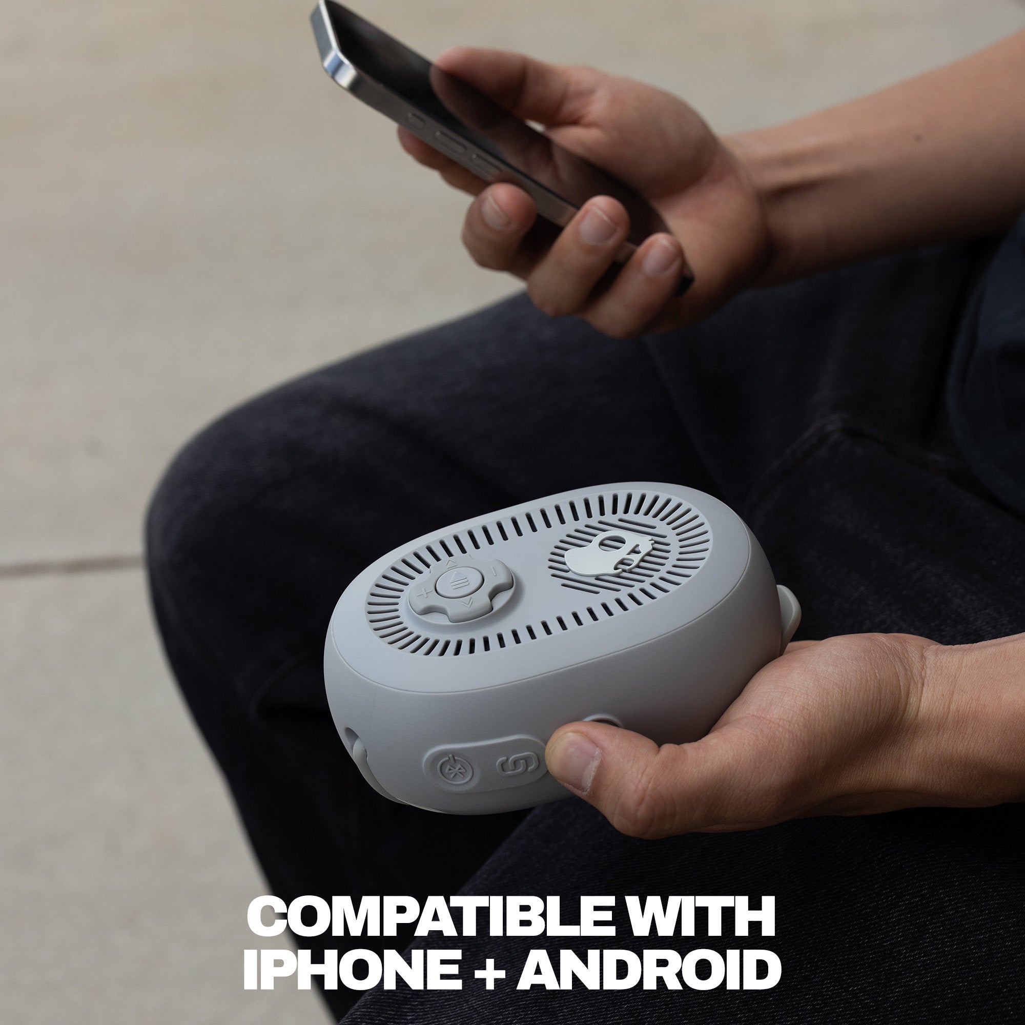 Person holding a small white device with a phone, sitting on a light-colored floor. | Cement