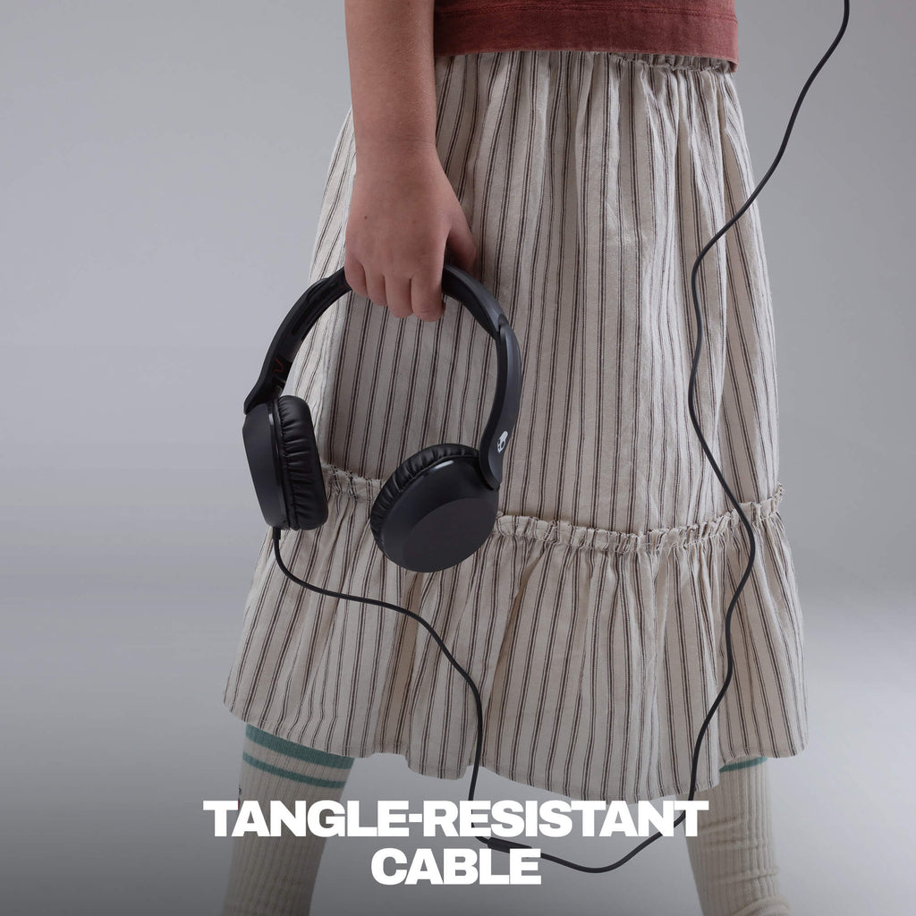 A child wearing a beige and white vertically striped skirt holds a pair of black over-ear headphones with a black tangle-resistant cable. The child is also wearing a rust-colored top and white socks. | True Black