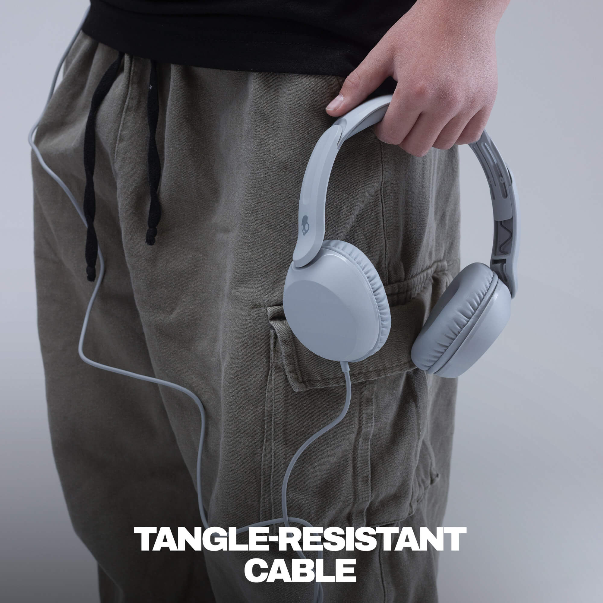 A person wearing loose olive cargo pants and a black top holds a pair of light gray over-ear headphones with a white cable tucked into the pants pocket. White text at the bottom reads 'TANGLE-RESISTANT CABLE'. | Cement
