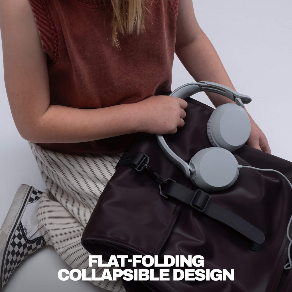 A person holds a dark brown flat-foldable backpack or laptop bag with gray over-ear headphones resting on top. The person wears a rust-colored sleeveless top and striped pants, seated against a light gray background. | Cement