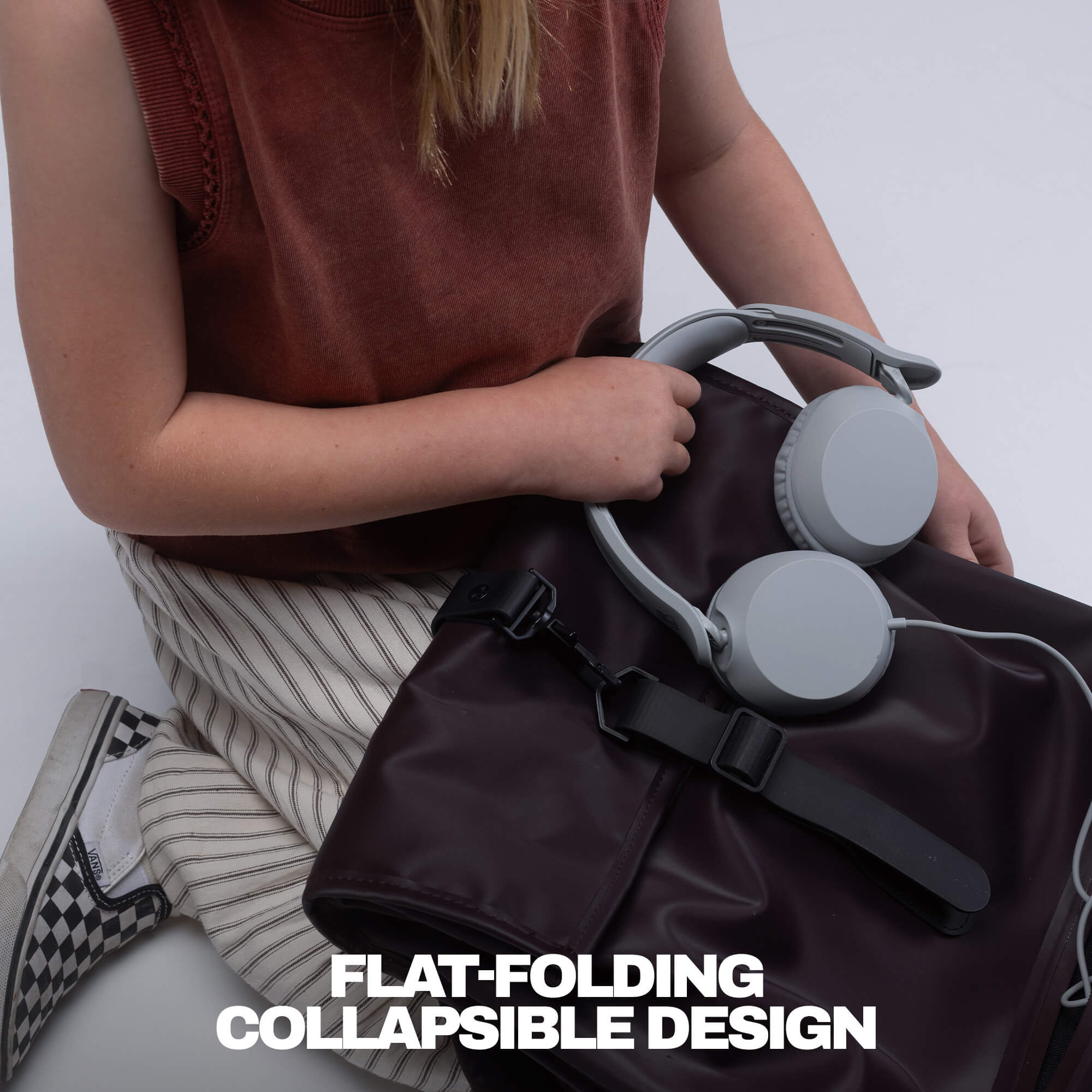 A person holds a dark brown flat-foldable backpack or laptop bag with gray over-ear headphones resting on top. The person wears a rust-colored sleeveless top and striped pants, seated against a light gray background. | Cement